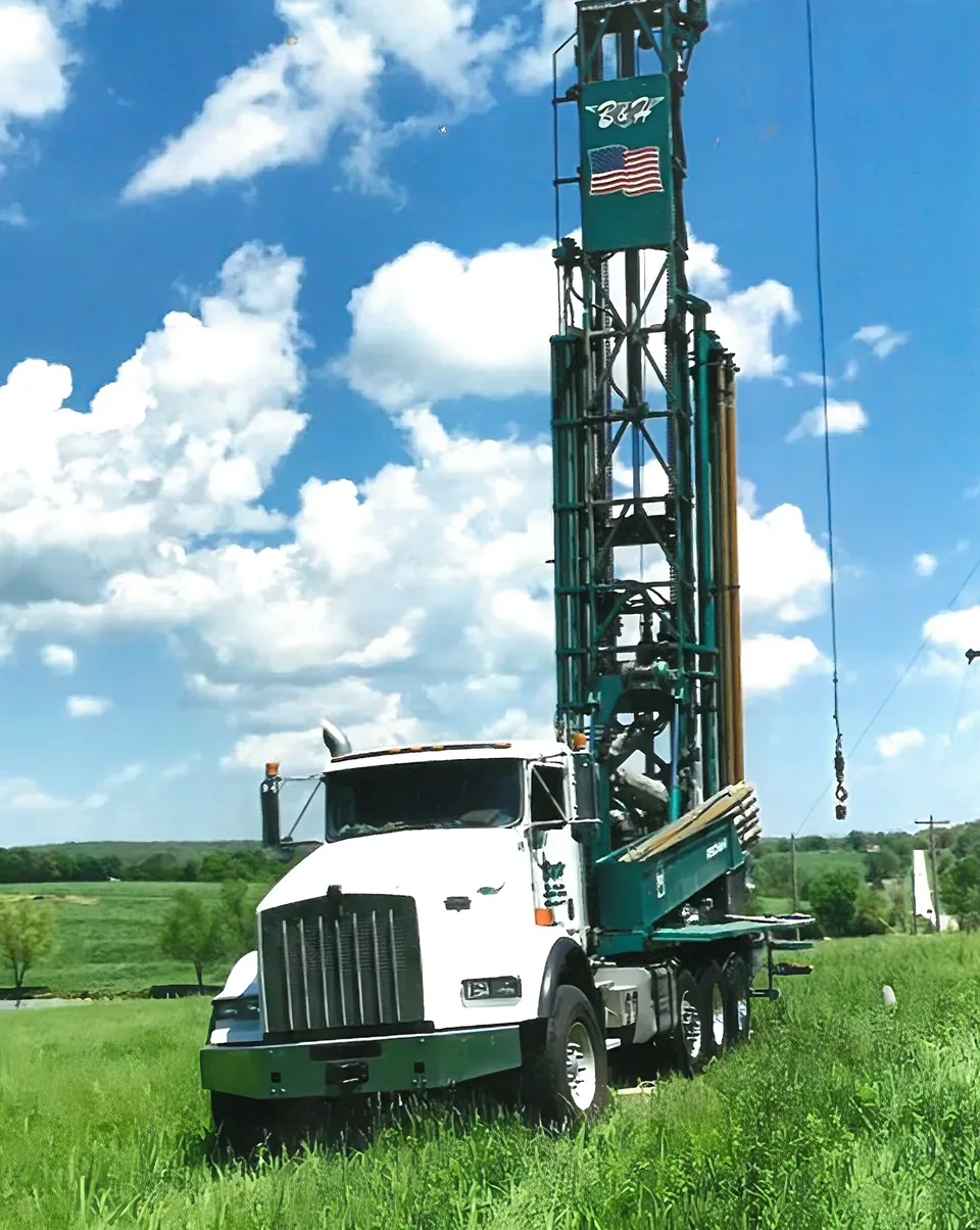 A B&H rotary drill rig with its mast raised, flying the American flag, drilling a water well in a Central Missouri field